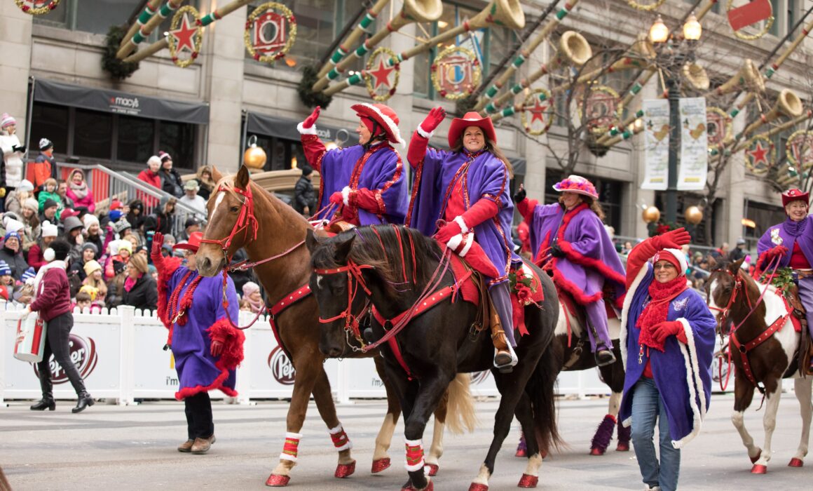 Several members (and their horses) of the Red Hats and Purple Chaps Equestrian Group marching in the 2016 Chicago Thanksgiving Parade. The ladies are adorned with red hats and purple coats.