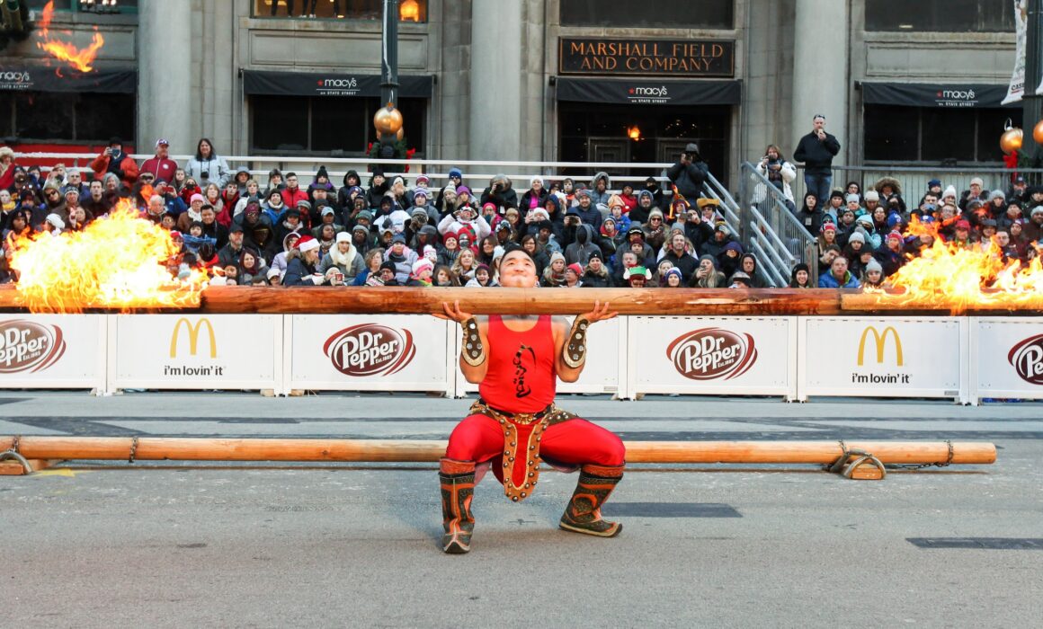 Strongman Tulga balances a giant log with flames on both ends in the TV Zone of the Chicago Thanksgiving Parade.
