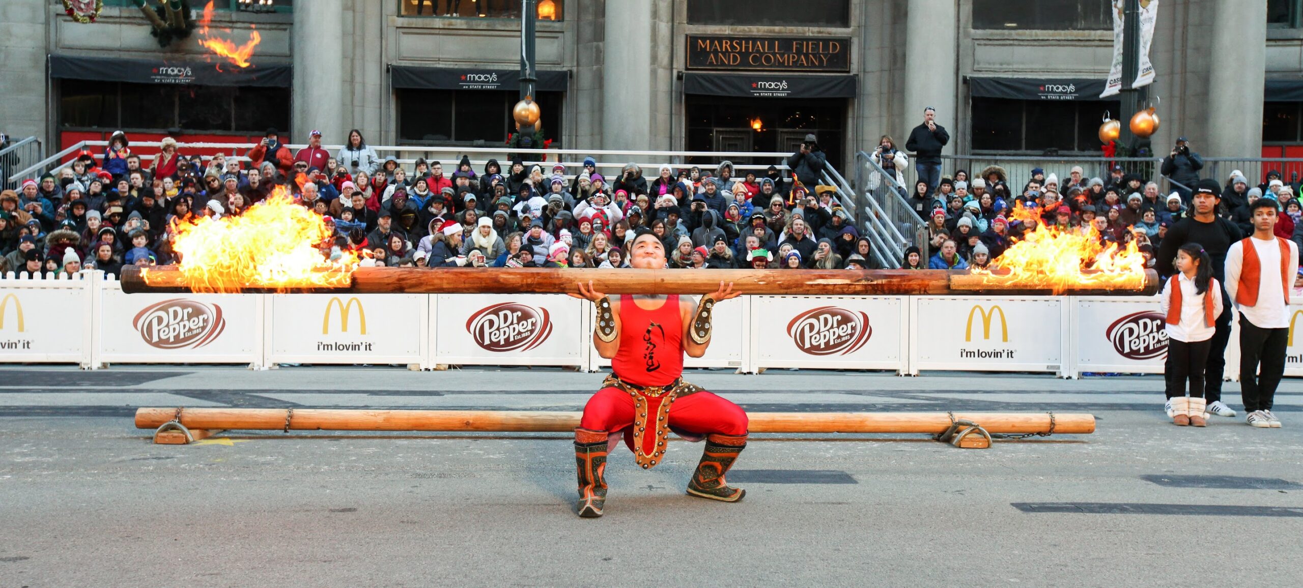 Strongman Tulga balances a giant log with flames on both ends in the TV Zone of the Chicago Thanksgiving Parade.